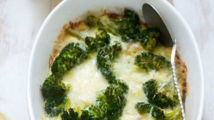 Ein Auflauf mit mangoldigem Gem&uuml;se, &uuml;berbacken mit K&auml;se, steht auf einem Tisch neben einer Sahneso&szlig;e und einem Holzbrett.