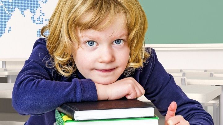 Ein Kind liegt l&auml;chelnd auf einem Stapel Schulb&uuml;cher, umgeben von einer Schulumgebung mit einer Weltkarte im Hintergrund.