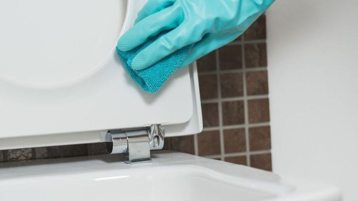 Eine Hand mit blauen Gummihandschuhen reinigt den Toilettendeckel, um Urinstein und Kalk zu entfernen.
