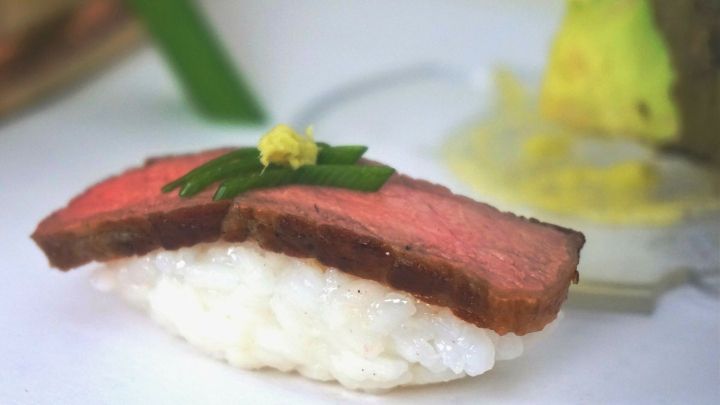 Ein St&uuml;ck Sushi liegt auf Reis, belegt mit zartem, rotem Fleisch, umgeben von frischen Zutaten in einem eleganten Ambiente.