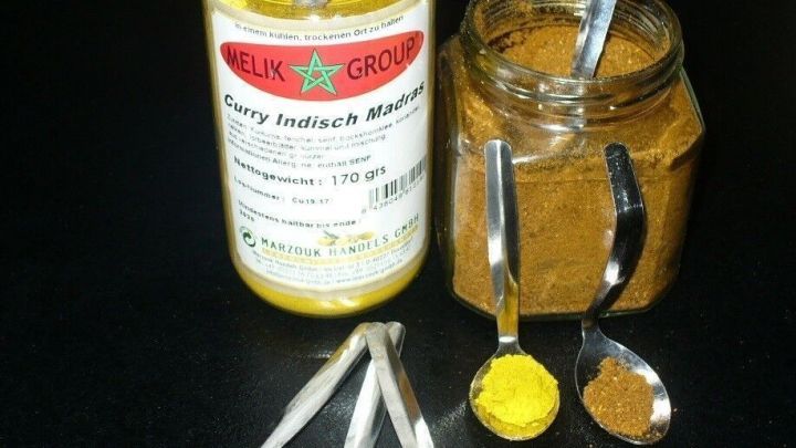 Ein Gew&uuml;rzportionierer aus kleinen Teel&ouml;ffeln f&uuml;llt bunte Gew&uuml;rze, w&auml;hrend zwei Gl&auml;ser mit verschiedenen Gew&uuml;rzmischungen daneben stehen.