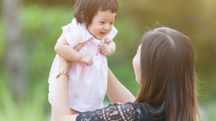 Eine Mutter hebt ihr Baby l&auml;chelnd in die Luft, w&auml;hrend sie im Sonnenlicht in einem gr&uuml;nen Park steht.