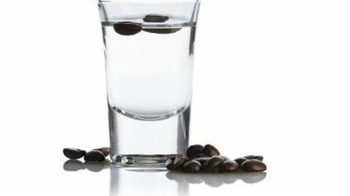 Ein klarer Sambuca-Schnapsglas enth&auml;lt treibende Kaffeebohnen, umgeben von verstreuten Bohnen auf einer wei&szlig;en Oberfl&auml;che.