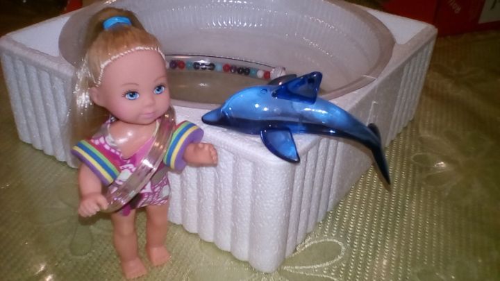 Eine kleine Puppenfigur steht neben einem selbstgemachten Planschbecken und h&auml;lt ein Spielzeugmesser, w&auml;hrend ein blauer Delfin schwimmt.