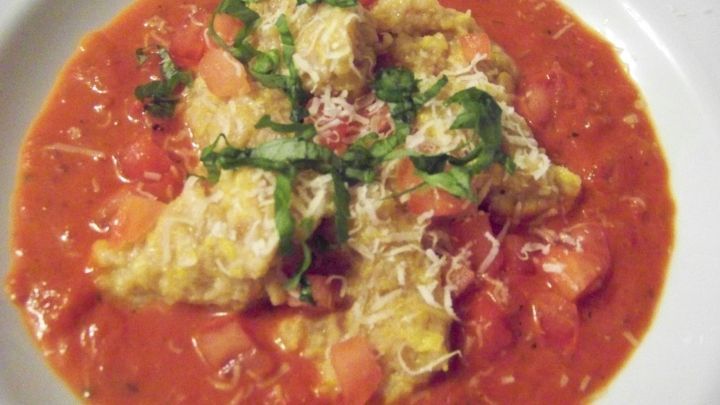 K&uuml;rbis-Malfatti liegen in einer tomatensaucenartigen Umgebung, garniert mit frischen Kr&auml;utern und K&auml;se.