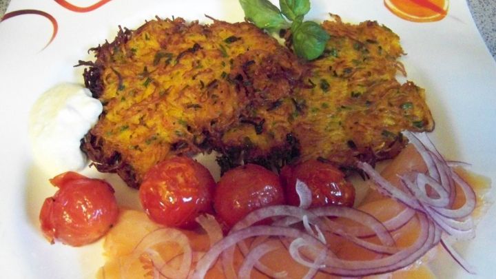 Knusprige Puffer vom K&uuml;rbis liegen auf einem Teller, umgeben von R&auml;ucherlachs, Zwiebeln und Kirschtomaten.