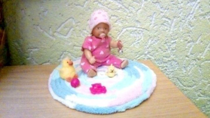 Eine Mini Babypuppe sitzt auf einer bunten Krabbeldecke und spielt mit einem gelben Stofftier in einem hellen Raum.