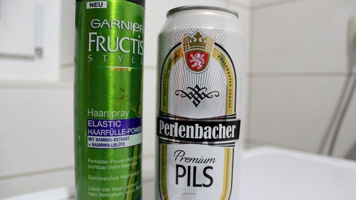 Eine Haarspray-Dose steht neben einer Bierdose, beide sind in einem Badezimmer auf einer wei&szlig;en Ablage platziert.