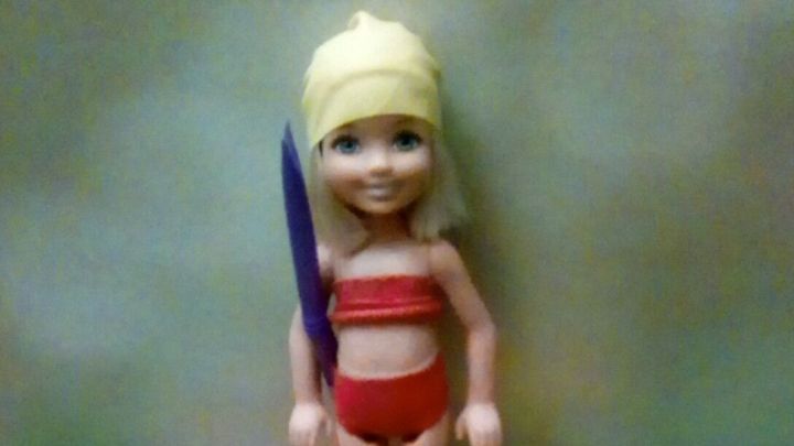 Eine kleine Puppe steht auf einer bunten Luftmatratze, neben ihr liegt eine Poolnudel und ein Ball.