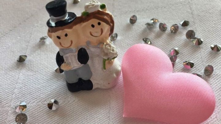 Eine Hochzeitsfigur steht neben einem pinken Herz, umgeben von glitzernden Dekorationen auf einem tisch&auml;hnlichen Untergrund.