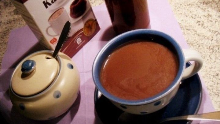 Ein tiefblauer Becher mit hei&szlig;em Kakao steht auf einem Tisch, umgeben von einem Zuckergef&auml;&szlig; und einer Packung Kakao.