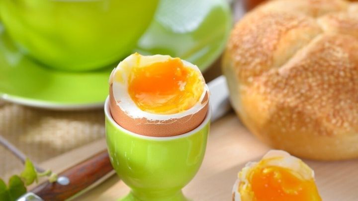 Ein perfekt gekochtes Fr&uuml;hst&uuml;cksei steht in einem gr&uuml;nen Eierbecher, umgeben von Marmelade, Br&ouml;tchen und einem Getr&auml;nk.