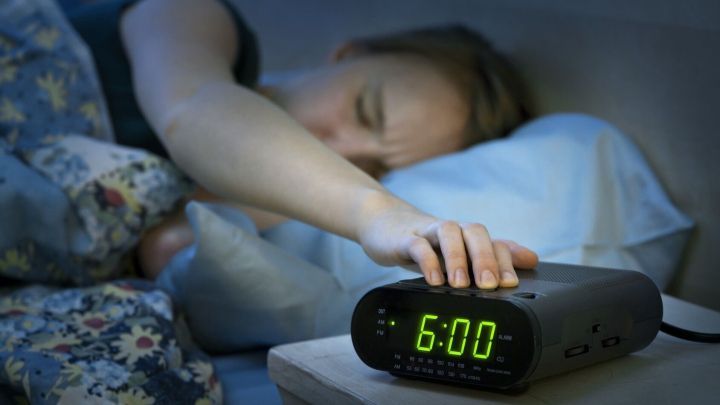Ein Wecker mit gr&uuml;nem Display zeigt 6:00 Uhr an, w&auml;hrend jemand im Bett im Dunkeln nach dem Einschaltknopf greift.