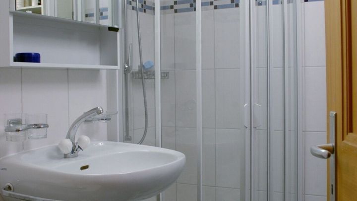 Eine Duschkabine steht in einem hellen Badezimmer mit wei&szlig;en Fliesen, w&auml;hrend das Waschbecken f&uuml;r die Reinigung genutzt wird.