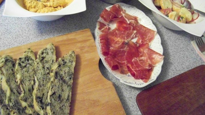 Ein dunkles Brot mit Mangold liegt auf einem Holzbrett, umgeben von verschiedenen Dips und Schinken auf einem Tisch.