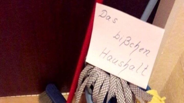 Besen und Wischmob liegen zusammen auf dem Boden, w&auml;hrend ein Schild mit &bdquo;Das bisschen Haushalt&ldquo; dar&uuml;ber h&auml;ngt.