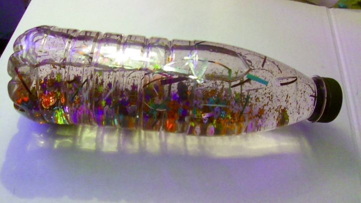 Eine Glitzerflasche steht auf einem Tisch, gef&uuml;llt mit buntem Glitzer und kleinen Dekorationen, die im Licht funkeln.