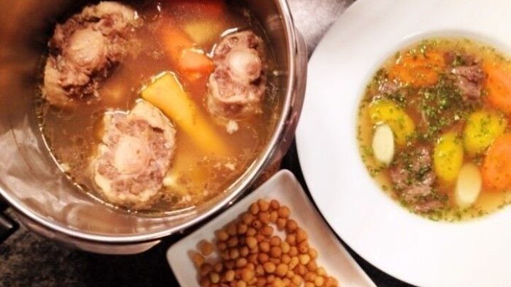 Eine dampfende Ochsenschleppsuppe befindet sich in einem Schnellkochtopf, w&auml;hrend eine servierfertige Portion mit Gem&uuml;se und Br&uuml;he auf einem Teller liegt. Daneben sind eingeweichte Kichererbsen zu sehen.