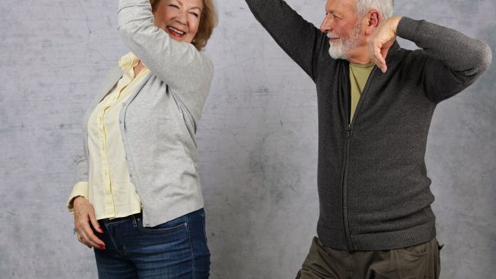 Ein &auml;lteres Paar tanzt ausgelassen in einem neutralen Raum, w&auml;hrend sie Freude und Lebenslust trotz Knieproblemen zeigen.