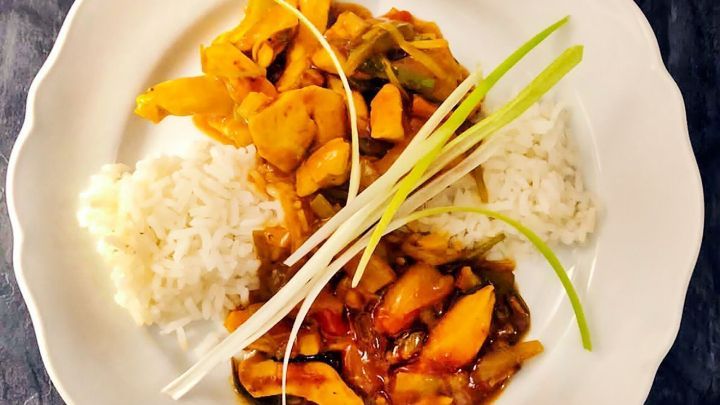 Ein Teller mit gebratenem H&uuml;hnchen in S&uuml;&szlig;-Sauer-So&szlig;e, umgeben von duftendem Reis und frischen Fr&uuml;hlingszwiebeln.