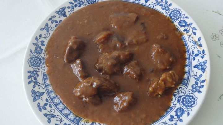 Ein Teller mit Saftgulasch zeigt zarte Fleischst&uuml;cke in dicker, dunkler Sauce auf einem blauen, gemusterten Teller.