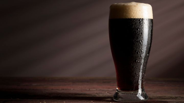 Ein mit Schaum gef&uuml;lltes Glas dunkles Bier steht auf einem groben Holztisch, von sanftem Licht beleuchtet.