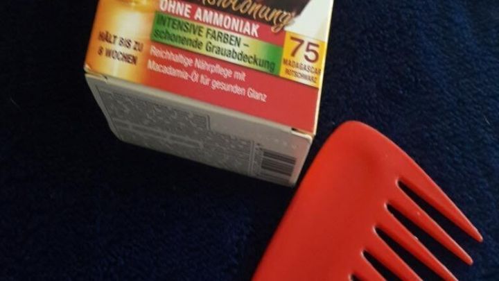 Eine rote Haarkamm liegt neben einer Haarf&auml;rbe-Box, w&auml;hrend die Verpackung eine Frau mit gl&auml;nzenden, gef&auml;rbten Haaren zeigt.