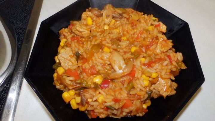 Ein bunter Reistopf mit Schweinefleisch liegt in einer schwarzen Schale und sieht appetitlich aus, umgeben von einer hellen Arbeitsfl&auml;che.