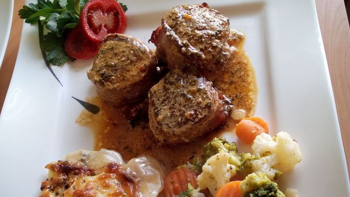 Filetscheiben vom Schwein im Speckmantel liegen auf einem Teller neben Kartoffelgratin und buntem Gem&uuml;se.