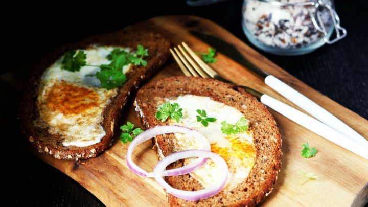 R&ouml;stbrot mit Ei liegt auf einem Holzbrett, garniert mit Zwiebeln und Kr&auml;utern, umgeben von einer dunklen Umgebung.