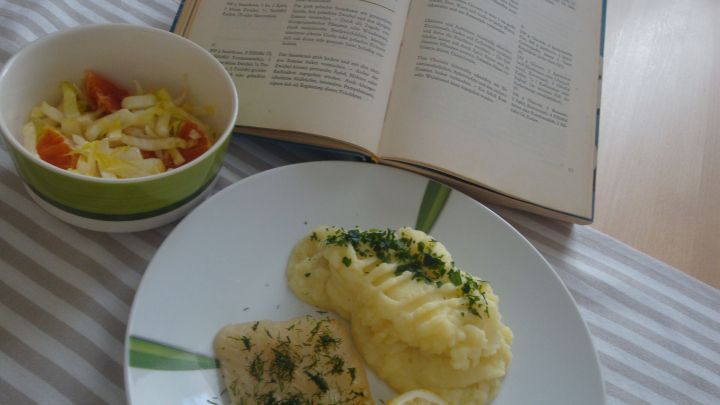 Ein Teller mit gefrorenem Heilbutt, serviert mit P&uuml;ree und einem Zitronenschnitz, neben einer Salatsch&uuml;ssel und einem offenen Kochbuch.