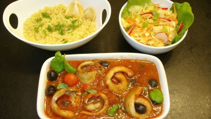 Gebackene Fischr&ouml;llchen in Tomatensauce liegen auf einem wei&szlig;en Teller, umgeben von einem Salat und Beilagen.