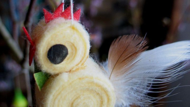 Ein buntes, handgefertigtes H&uuml;hnchen baumelt an einem Zweig, umgeben von bl&uuml;henden &Auml;sten und Fr&uuml;hlingsdeko.