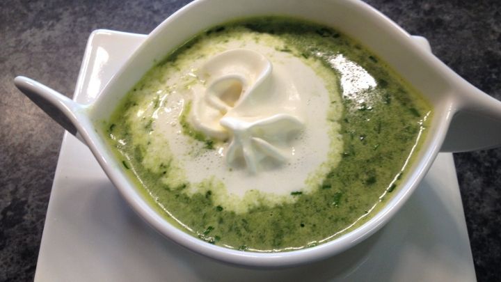 Eine Sch&uuml;ssel mit B&auml;rlauchcremesuppe, garniert mit Sahne, steht auf einem wei&szlig;en Teller in einer dunklen Umgebung.