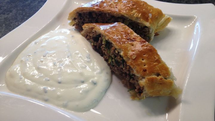 Goldbrauner Hackstrudel aus Bl&auml;tterteig liegt appetitlich auf einem wei&szlig;en Teller neben cremigem Dip.