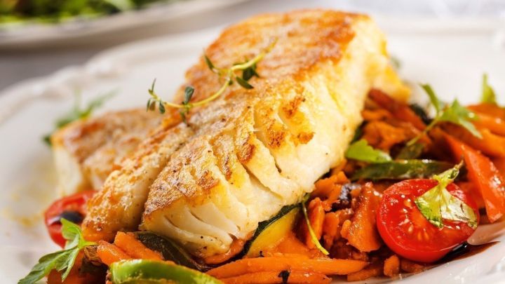 Goldgelber Fisch liegt auf einem frischen Gem&uuml;sebett aus Karotten und Tomaten, serviert in einladender Umgebung.