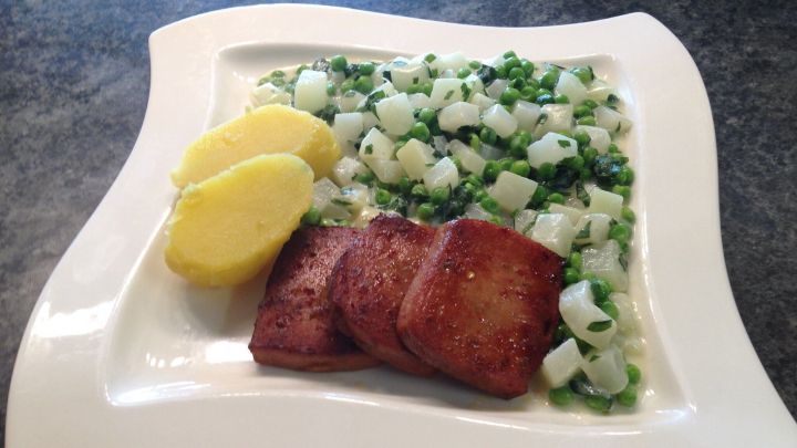 Kohlrabi und Erbsen sind auf einem wei&szlig;en Teller angerichtet, begleitet von zwei Kartoffeln, auf einem dunklen Tisch.