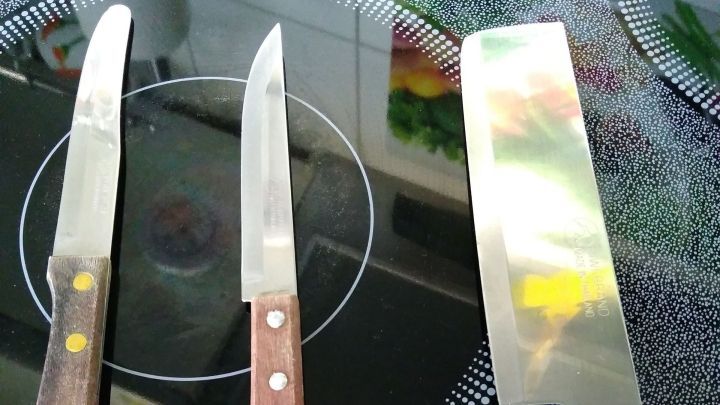 Drei scharfe Messer mit Holzgriffen liegen auf einer schwarzen Herdplatte, bereit f&uuml;r das Schneiden von Zutaten.