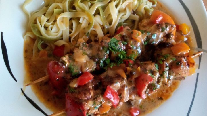 Saftige H&auml;hnchenspie&szlig;e liegen auf einem wei&szlig;en Teller, serviert mit Nudeln und buntem Gem&uuml;se in einer w&uuml;rzigen So&szlig;e.