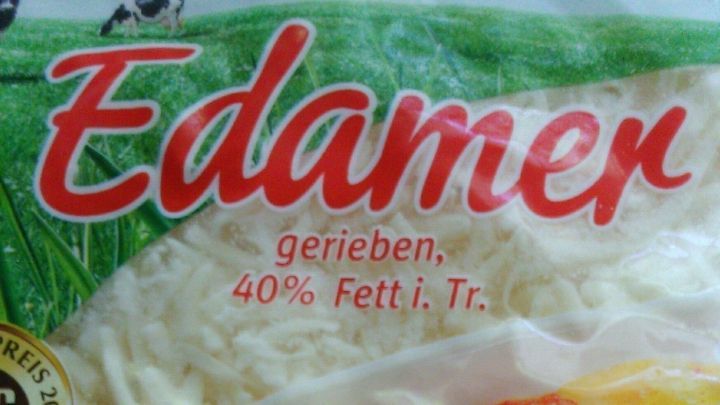 Eine Verpackung mit geriebenem Edamer-K&auml;se, die auf einem Tisch liegt, umgeben von buntem Gem&uuml;se.