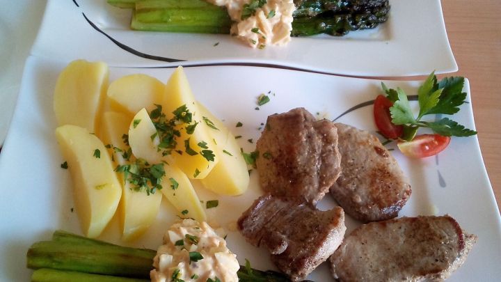 Gr&uuml;ner Spargel und Schweinefilets liegen auf einem Teller, umgeben von Kartoffeln und Bohnencreme in einem modernen Essbereich.