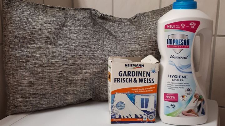 Ein graues Kissen liegt auf einer wei&szlig;en Oberfl&auml;che, daneben steht ein Waschmittel und eine Packung f&uuml;r Gardinen.