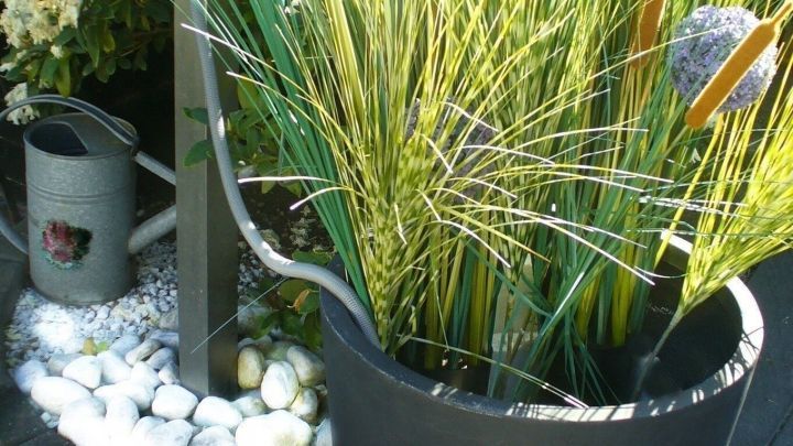 Ein gro&szlig;er schwarzer Topf mit k&uuml;nstlichen Gr&auml;sern steht auf einem Gehweg, umgeben von Kies und wei&szlig;en Steinen im Garten.