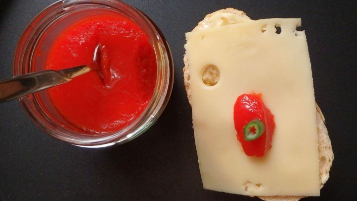 Ein Glas mit rotem Paprikagelee steht neben einer Scheibe Brot, belegt mit K&auml;se und einer Chili.