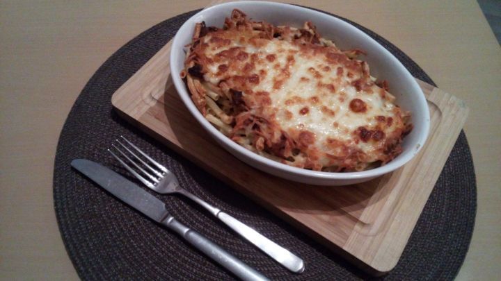 Ein gebackenes Nudelgericht mit w&uuml;rziger Hackfleischso&szlig;e in einer ovalen Schale auf einem Holzbrett, umgeben von Besteck.