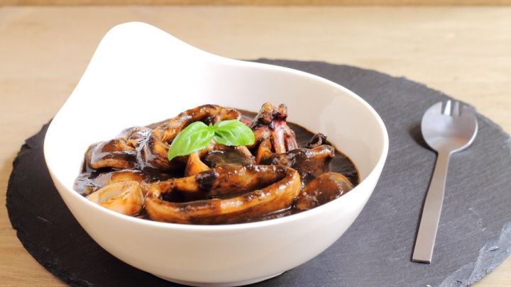 Ein wei&szlig;er Teller mit calamares a la cazuela, gef&uuml;llt mit Tintenfisch und einer Kr&auml;uterdekoration, auf einem schwarzen Untersetzer.