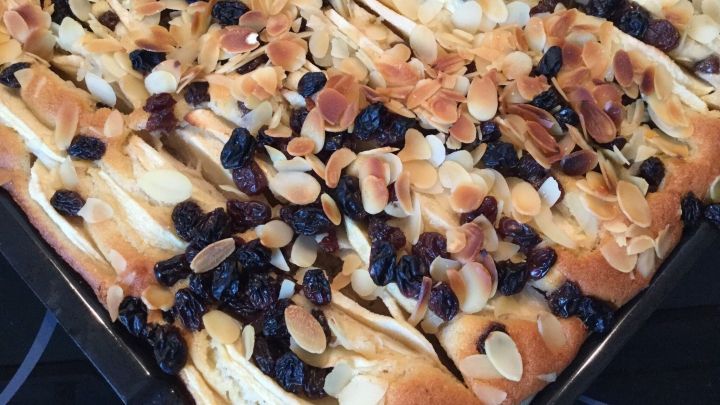 Ein goldbrauner Apfel-Blechkuchen liegt auf einem Blech, garniert mit Mandelsplittern und Rosinen in einer K&uuml;chenumgebung.