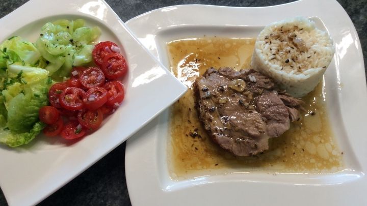 Ein saftiger Schweinsbraten liegt auf einem Teller mit So&szlig;e, begleitet von Reis und einem frischen Salat.