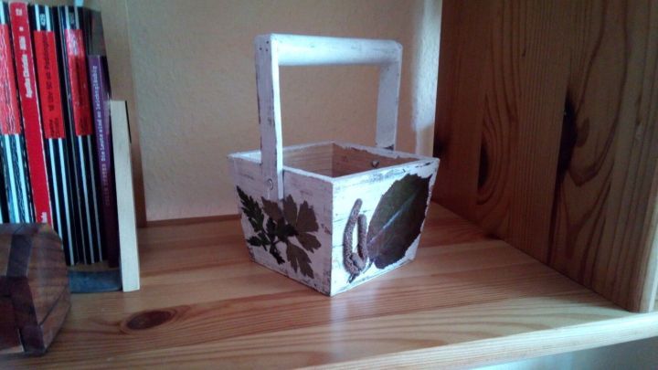 Ein kleiner, dekorativer Korb aus Papier steht auf einem Holzregal, verziert mit getrockneten Blumen und Bl&auml;ttern.