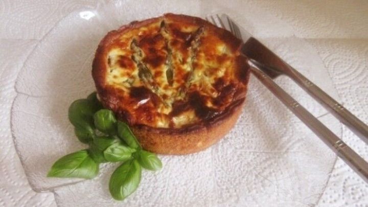Eine runde Spargel-Quiche auf einem eleganten Glas-Teller, garniert mit frischem Basilikum und Besteck daneben.